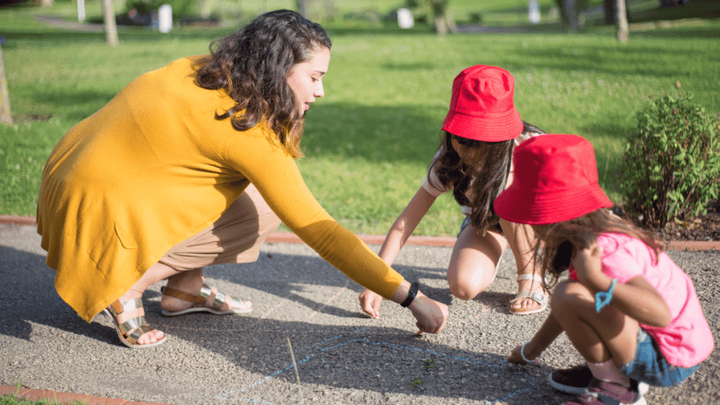Nannytax | Nanny recognition week Nanny playing with 2 small children in red hats, drawing on the ground with coloured chalk, with a grassy park behind them.